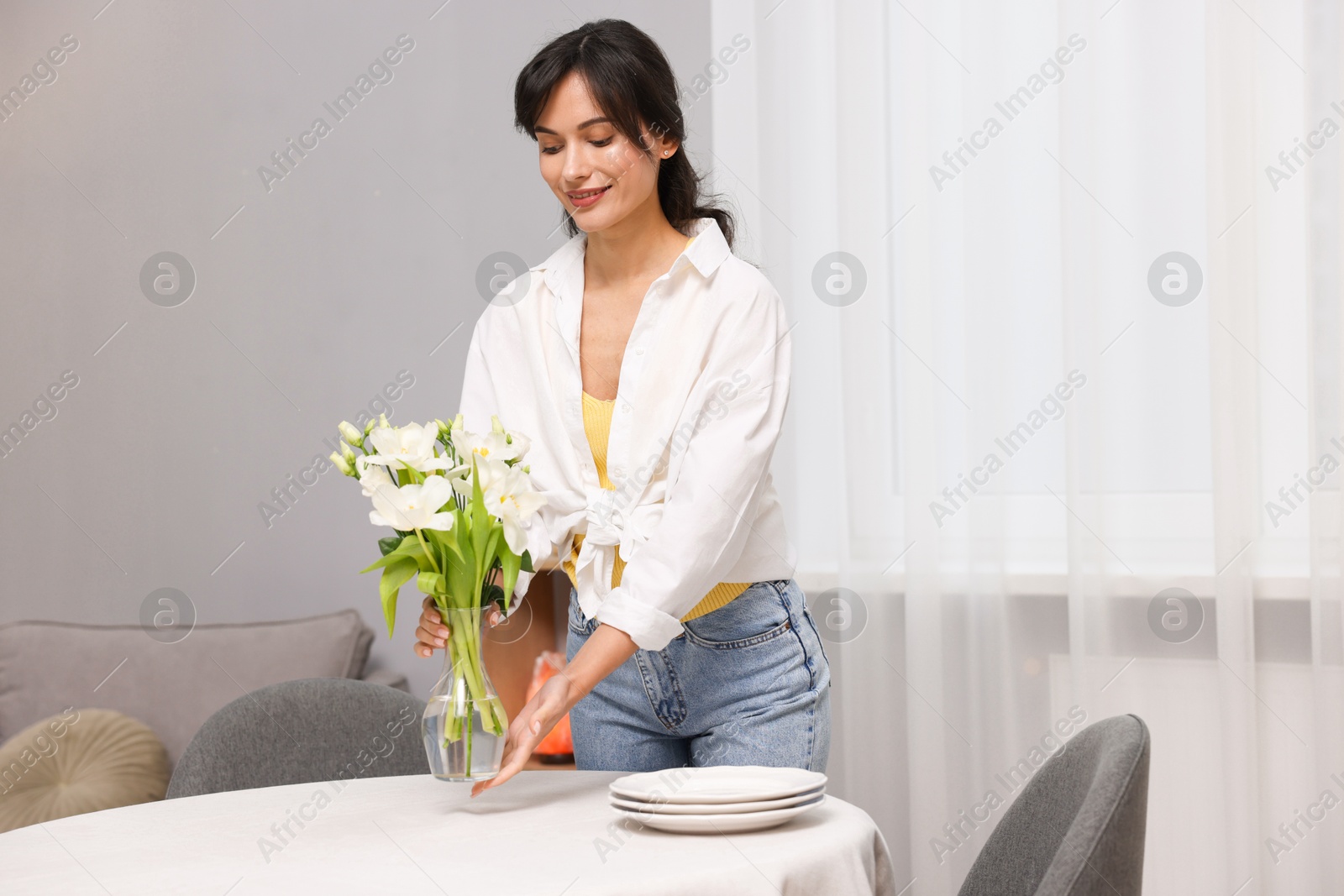 Photo of Young woman putting vase with flowers on table with white tablecloth at home