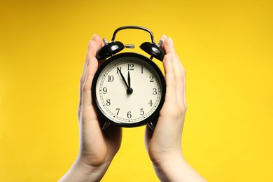 Photo of Woman with black alarm clock on yellow background, closeup