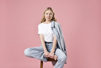 Beautiful young woman in stylish jeans sitting on stool against pink background