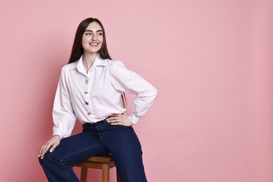 Photo of Smiling woman in stylish jeans sitting on stool against pink background. Space for text