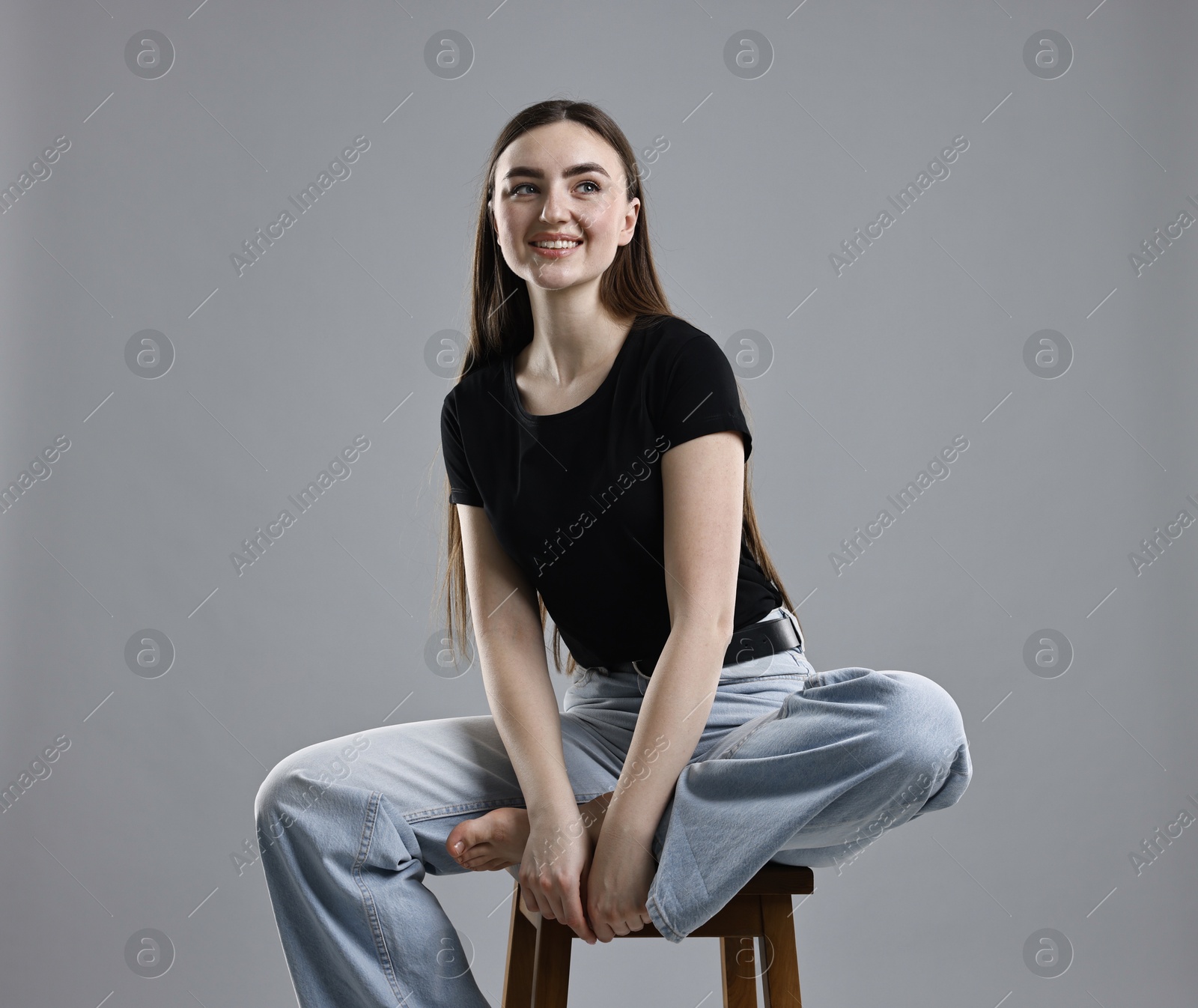 Photo of Smiling woman in stylish jeans sitting on stool against grey background