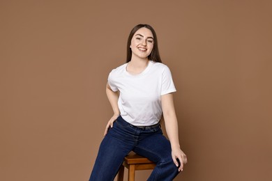 Photo of Smiling woman in stylish jeans sitting on stool against brown background