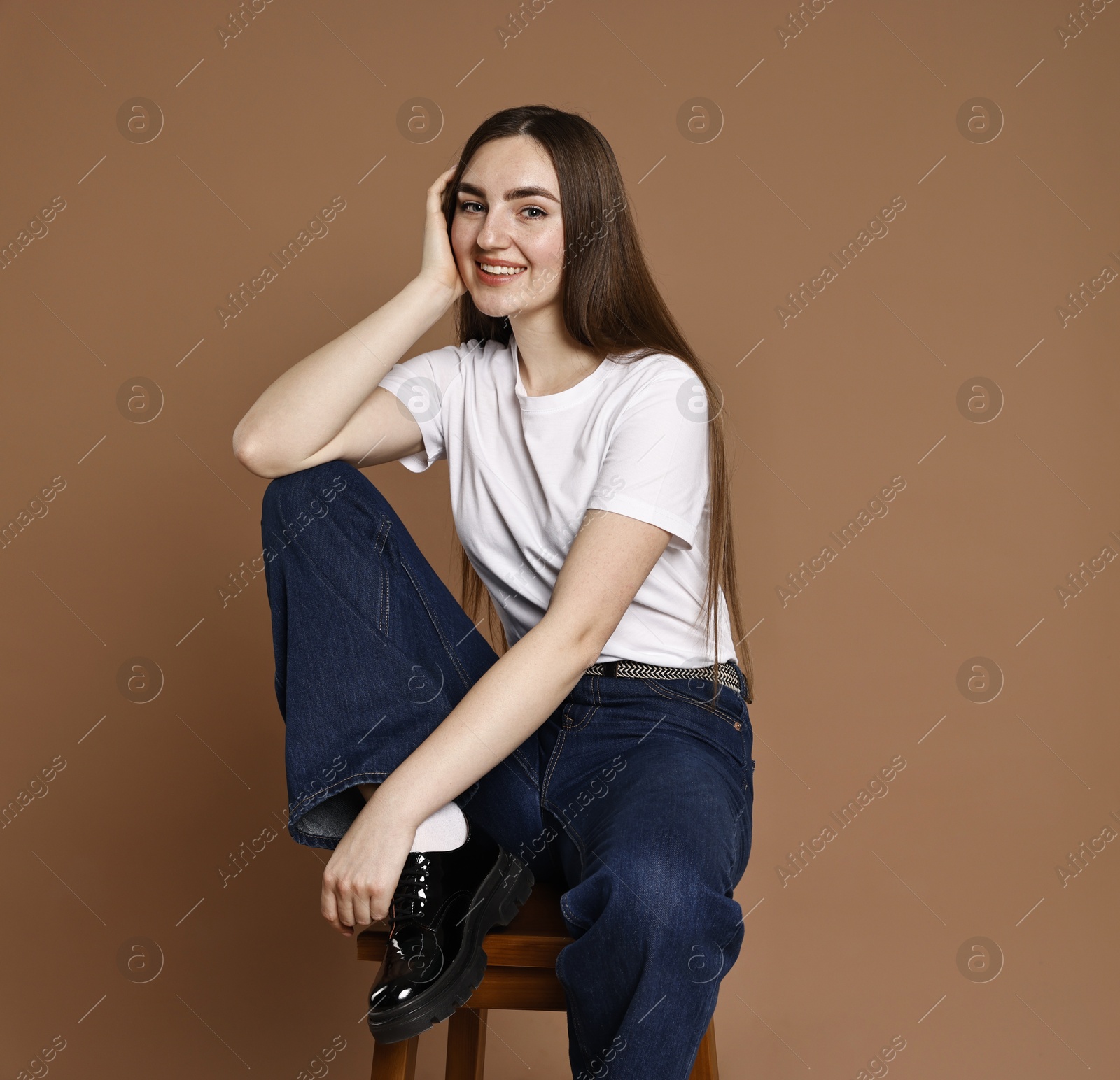 Photo of Smiling woman in stylish jeans sitting on stool against brown background. Space for text