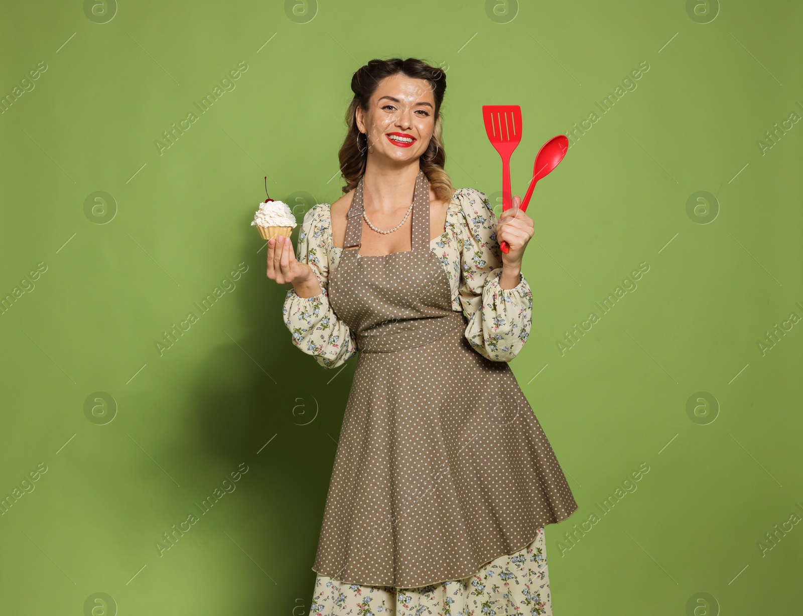 Photo of Happy woman with cupcake and cooking utensils on green background. Pin-up vibe portrait