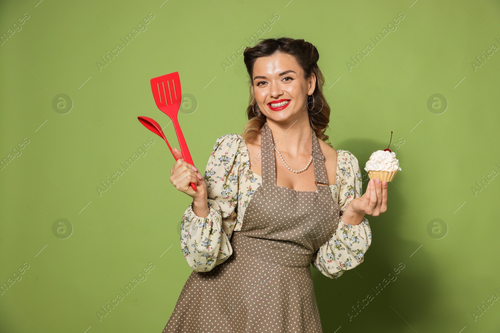 Photo of Happy woman with cupcake and cooking utensils on green background. Pin-up vibe portrait