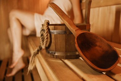 Photo of Bucket with ladle and woman on wooden bench in sauna, selective focus