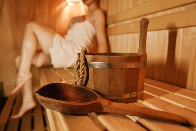 Photo of Bucket with ladle and woman on wooden bench in sauna, selective focus