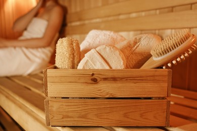 Crate with bath accessories and woman on wooden bench in sauna, selective focus