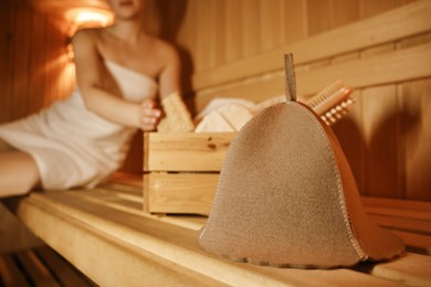 Woman with bath accessories and felt wool hat on wooden bench in sauna, selective focus