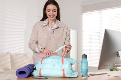 Photo of Happy woman with gym bag, fitness mat and water bottle at desk indoors