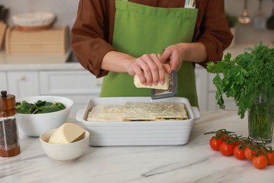 Photo of Woman grating cheese onto spinach lasagna at marble table indoors, closeup