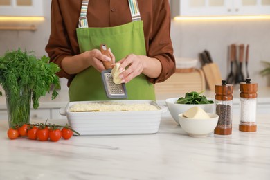 Photo of Woman grating cheese onto spinach lasagna at marble table indoors, closeup