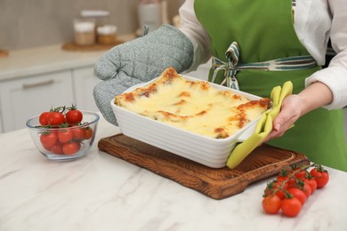 Photo of Woman putting baking dish with tasty lasagna onto white marble table indoors, closeup