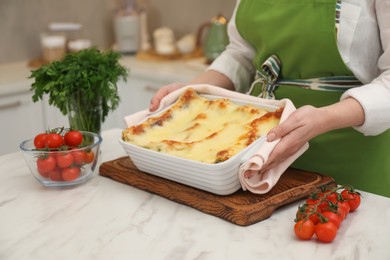 Photo of Woman putting baking dish with tasty lasagna onto white marble table indoors, closeup