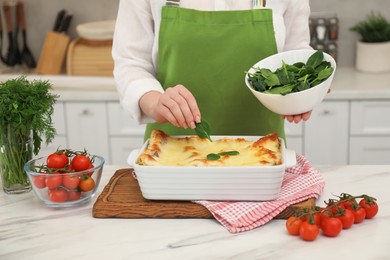 Photo of Woman putting spinach onto lasagna at white marble table in kitchen, closeup