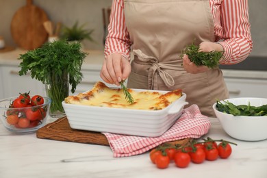 Photo of Woman putting rosemary onto lasagna at white marble table in kitchen, closeup