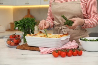 Photo of Woman putting rosemary onto lasagna at white marble table in kitchen, closeup