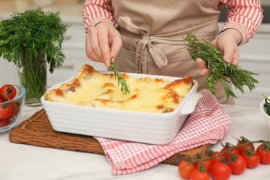 Photo of Woman putting rosemary onto lasagna at white marble table indoors, closeup