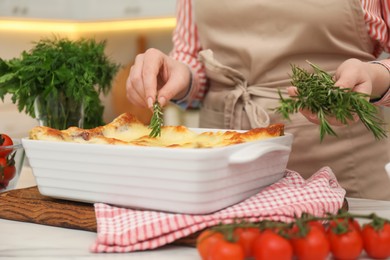 Photo of Woman putting rosemary onto tasty lasagna at white table indoors, closeup