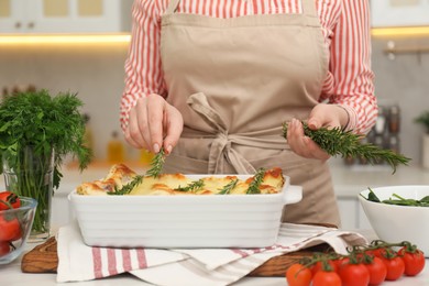 Photo of Woman putting rosemary onto lasagna at white table in kitchen, closeup