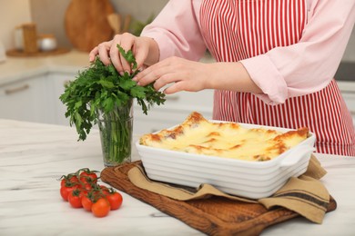 Photo of Woman taking parsley from glass near delicious lasagna at white marble table indoors, closeup