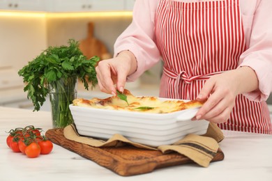 Photo of Woman putting parsley onto lasagna at white marble table indoors, closeup