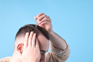 Baldness problem. Man applying serum onto hairline on light blue background, closeup