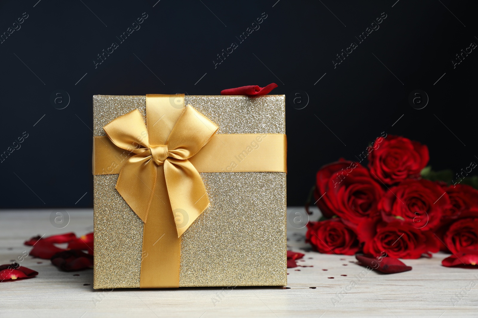 Photo of Beautiful gift box, rose flowers, petals and confetti on white wooden table against dark background, closeup. Romantic present