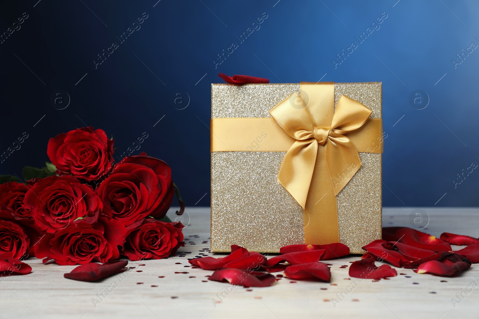 Photo of Beautiful gift box, rose flowers, petals and confetti on white wooden table against blue background, closeup. Romantic present