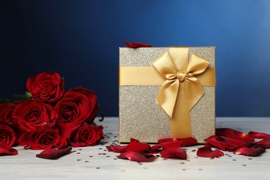 Beautiful gift box, rose flowers, petals and confetti on white wooden table against blue background, closeup. Romantic present