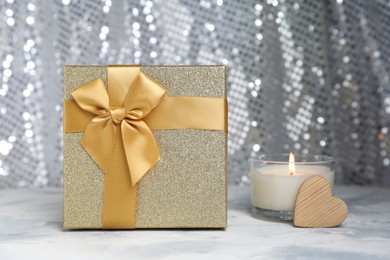 Beautiful gift box, wooden heart and burning candle on table against silver glitter background. Romantic present