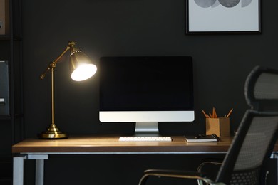 Photo of Comfortable workplace with computer and lamp on wooden desk at home office in evening