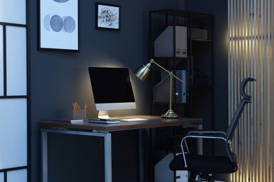 Photo of Comfortable workplace with computer and lamp on wooden desk at home office in evening