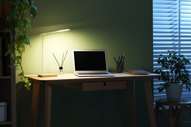 Photo of Comfortable workplace with laptop and lamp on wooden desk at home office in evening