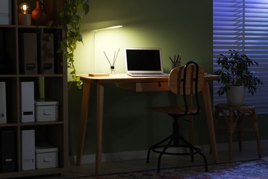 Photo of Comfortable workplace with laptop and lamp on wooden desk at home office in evening