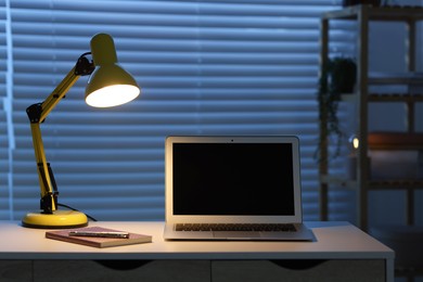 Photo of Comfortable workplace with laptop and lamp on desk at home office in evening