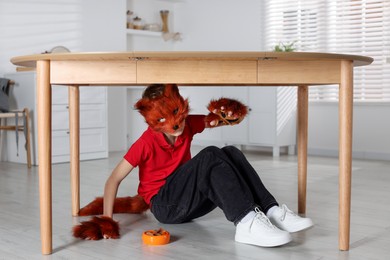 Quadrobics. Boy wearing fox mask, tail and gloves with feeding bowl under table indoors