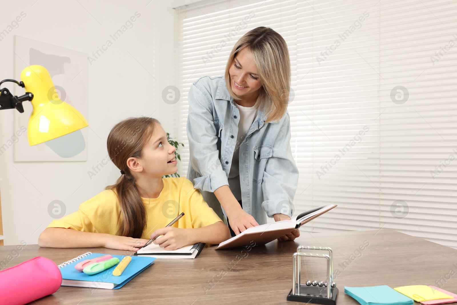 Photo of Smiling mother helping her daughter with homework at table indoors