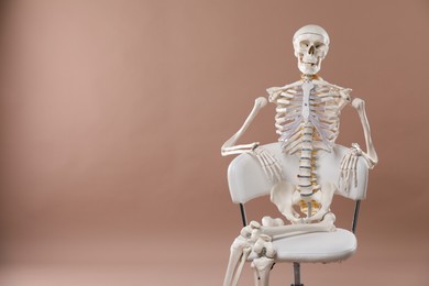 Artificial human skeleton model sitting on chair against light brown background, space for text