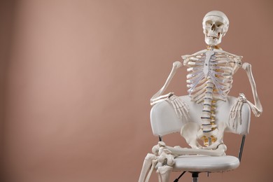 Artificial human skeleton model sitting on chair against light brown background, space for text