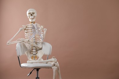 Artificial human skeleton model sitting on chair against light brown background, space for text