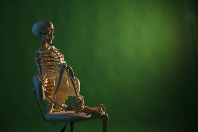 Artificial human skeleton model sitting on chair against green background, space for text