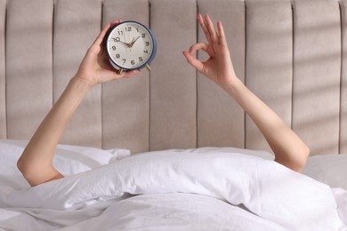 Woman with alarm clock showing ok gesture in bed at home, closeup. Good morning