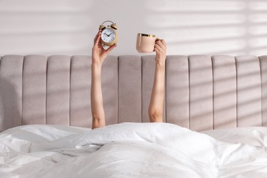 Woman with alarm clock and cup in bed at home, closeup. Good morning