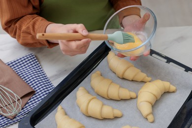 Photo of Woman brushing egg wash onto raw croissants at white marble table, closeup