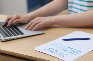 Photo of Student using laptop at wooden table, focus on scholarship application form and pen