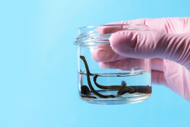 Photo of Woman holding glass jar with medicinal leeches on light blue background, closeup. Space for text