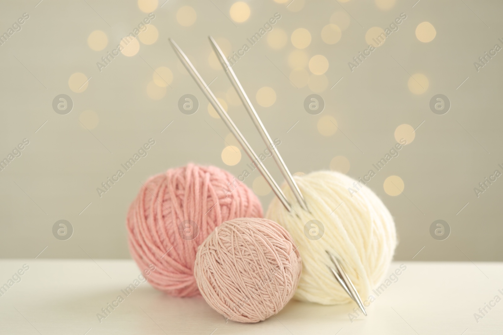 Photo of Balls of colorful yarn and knitting needles on white table against grey background with blurred lights, bokeh effect