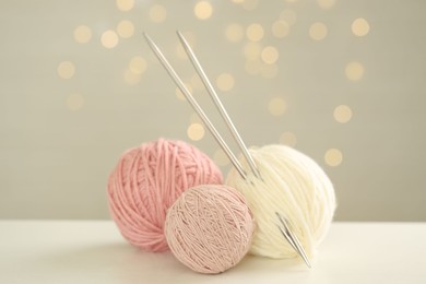 Balls of colorful yarn and knitting needles on white table against grey background with blurred lights, bokeh effect