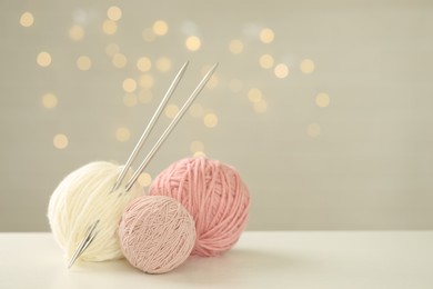 Balls of colorful yarn and knitting needles on white table against grey background with blurred lights, space for text. Bokeh effect